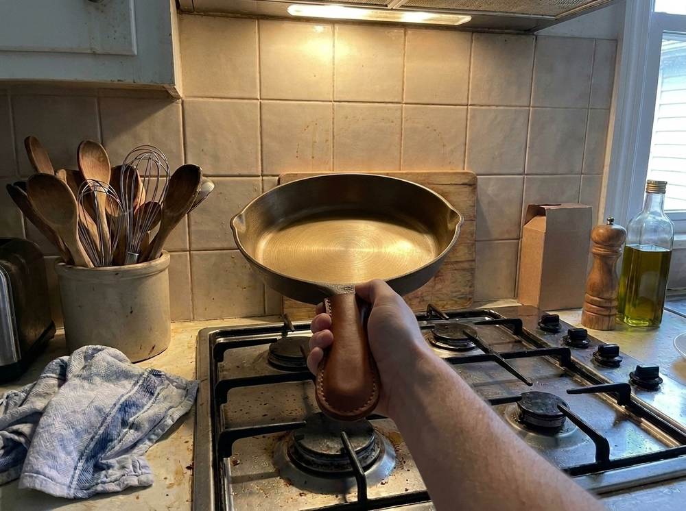 One hand holding the iLume up off the stove by its leather-covered handle, the stitched saddle-brown cover facing the camera.