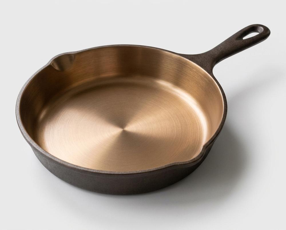 The iLume cast iron skillet — polished bronze interior, dark sand-cast exterior, pour spout and teardrop hanging hole visible.