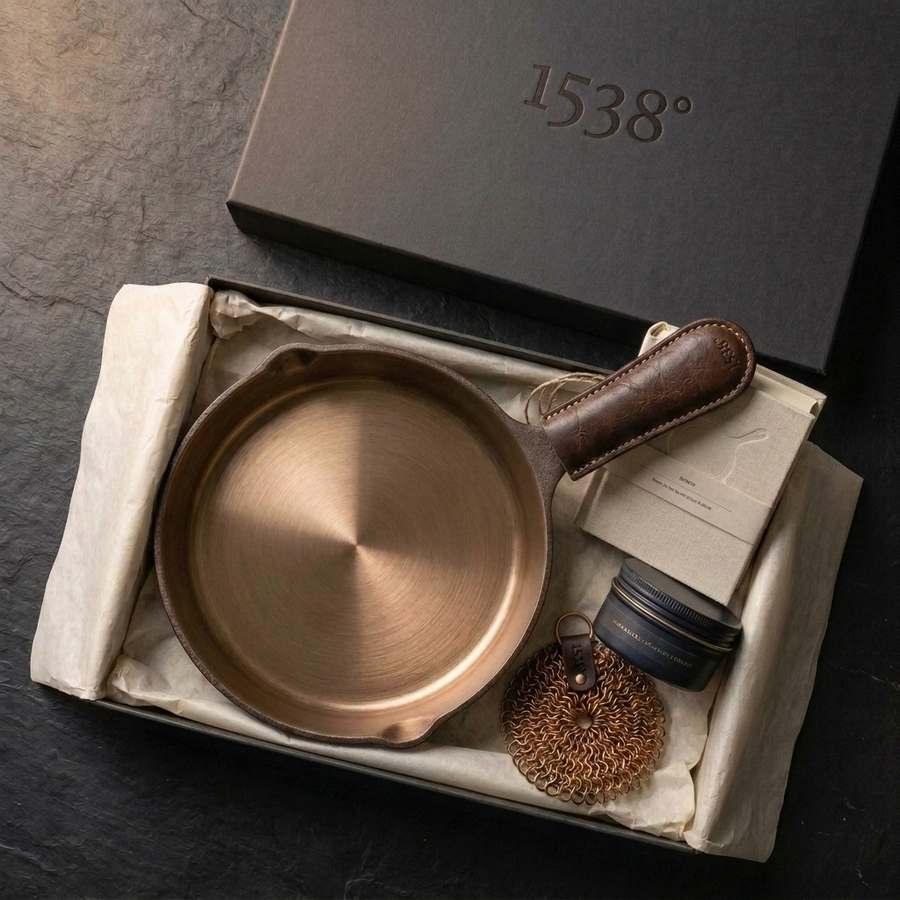 The iLume box open on a dark counter: the bronze-interior skillet wearing its leather handle sleeve, a 'The First 30 Days' starter card, and a tin of seasoning oil nested in tissue paper, with the embossed lid resting behind.